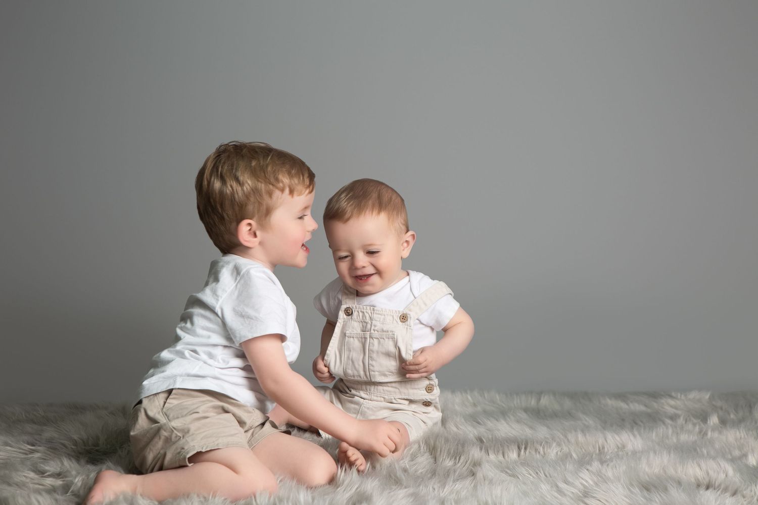 Two young boys in white shirts sitting on a fluffy gray rug, smiling and interacting with each other.