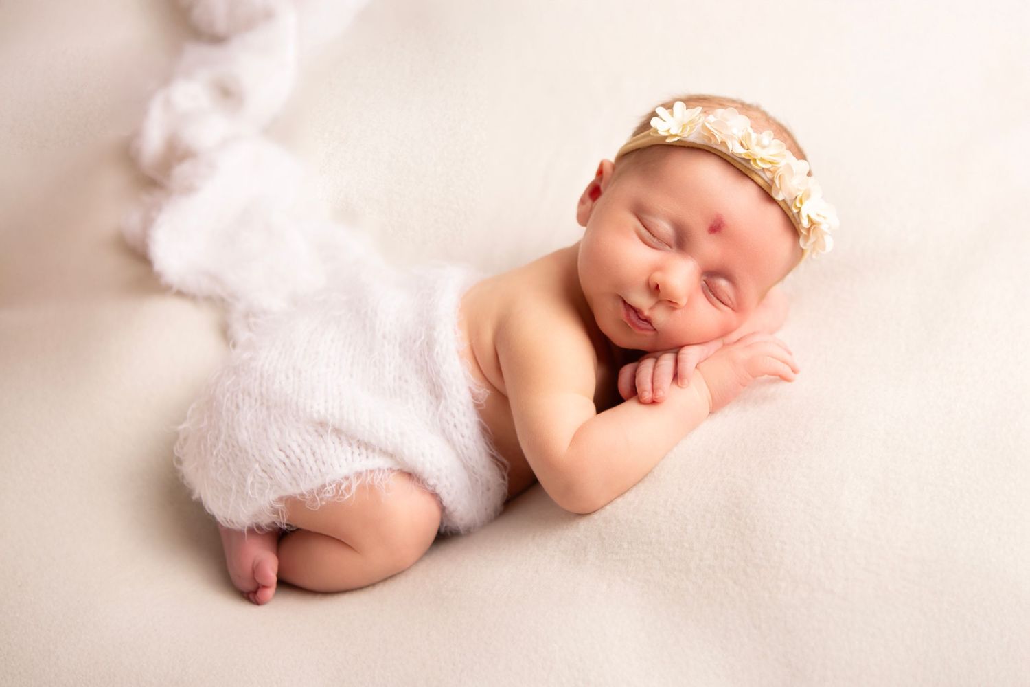 Peaceful newborn baby sleeping wrapped in white fluffy fabric with a delicate floral headband on a cream background.