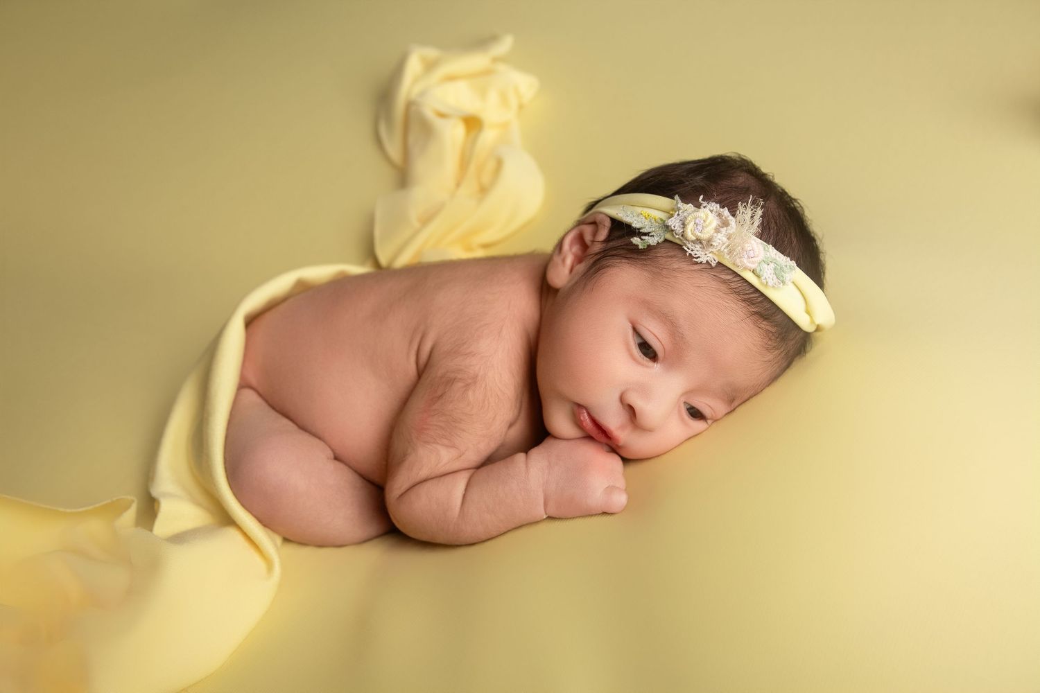 Newborn baby girl wrapped in yellow fabric with floral headband, peacefully posed on soft yellow background.