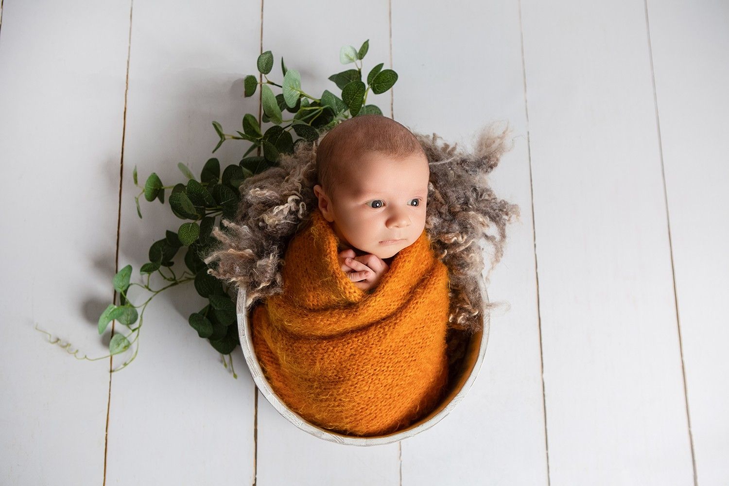 Newborn baby swaddled in orange wrap, nestled in wooden bowl with fur and eucalyptus, on white wood floor.