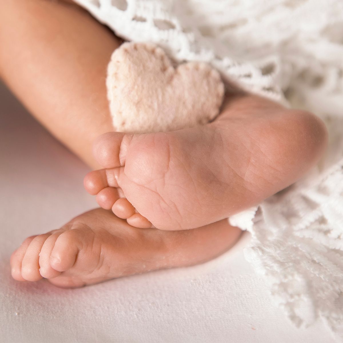 Close-up of tiny newborn baby feet with a small plush heart toy on a soft white lace blanket.