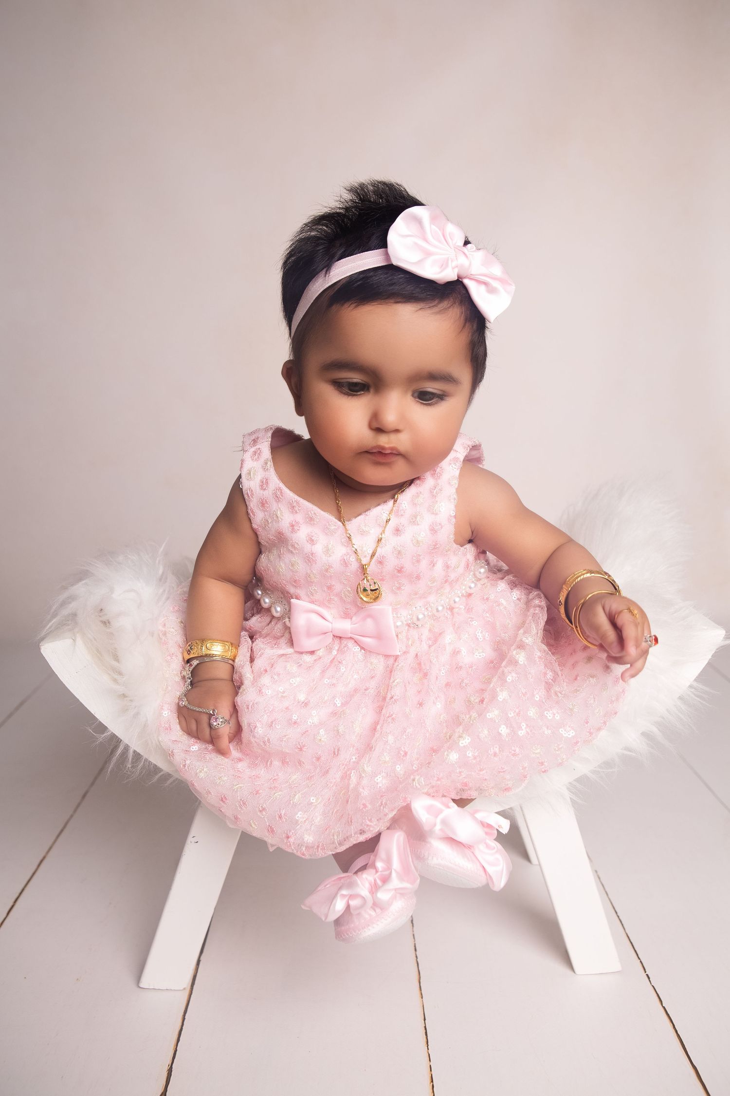 Adorable baby girl in pink polka dot dress with bow headband and gold jewelry sitting on white stool.