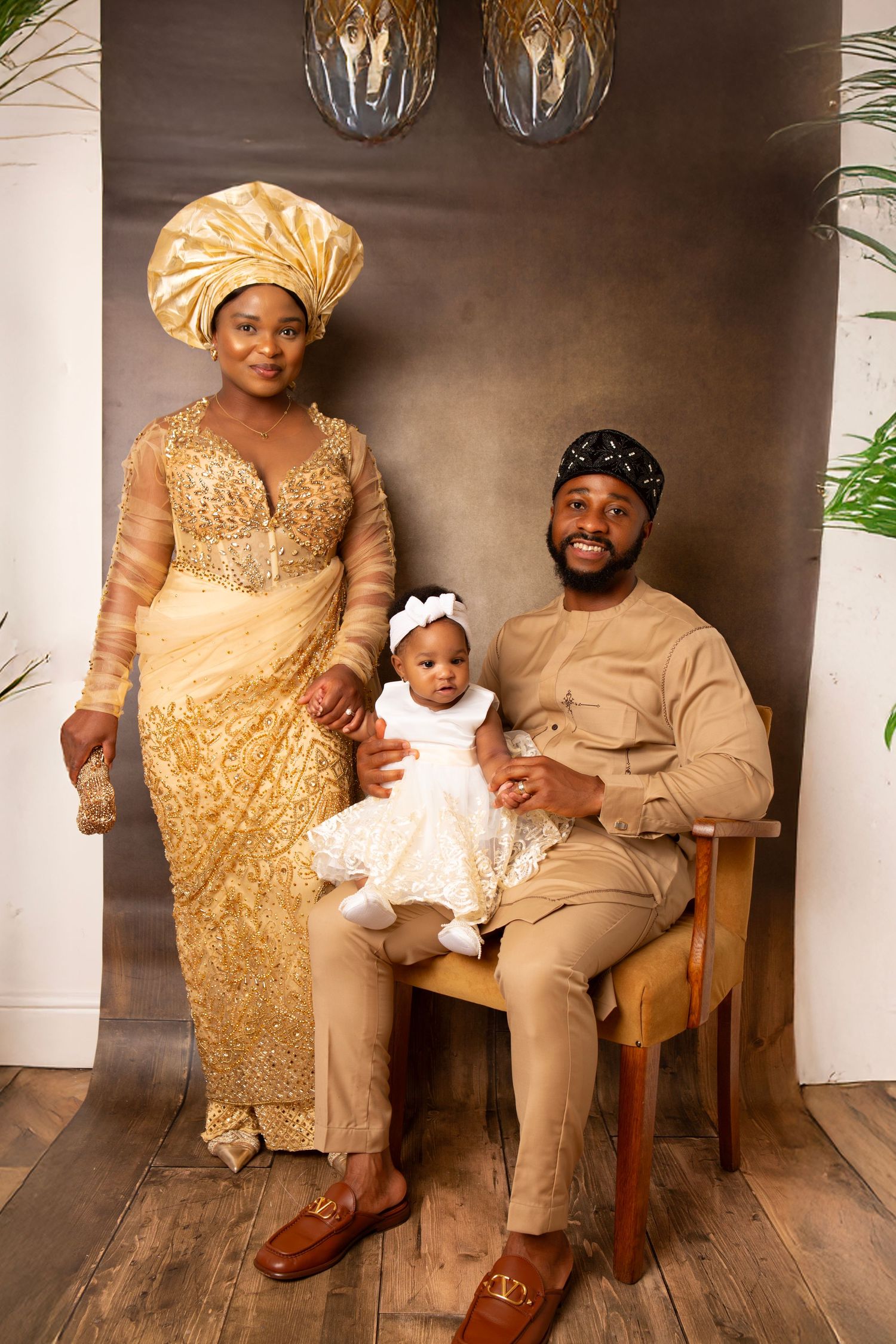 Nigerian family in traditional gold attire with baby in white dress posing for a formal portrait photo.