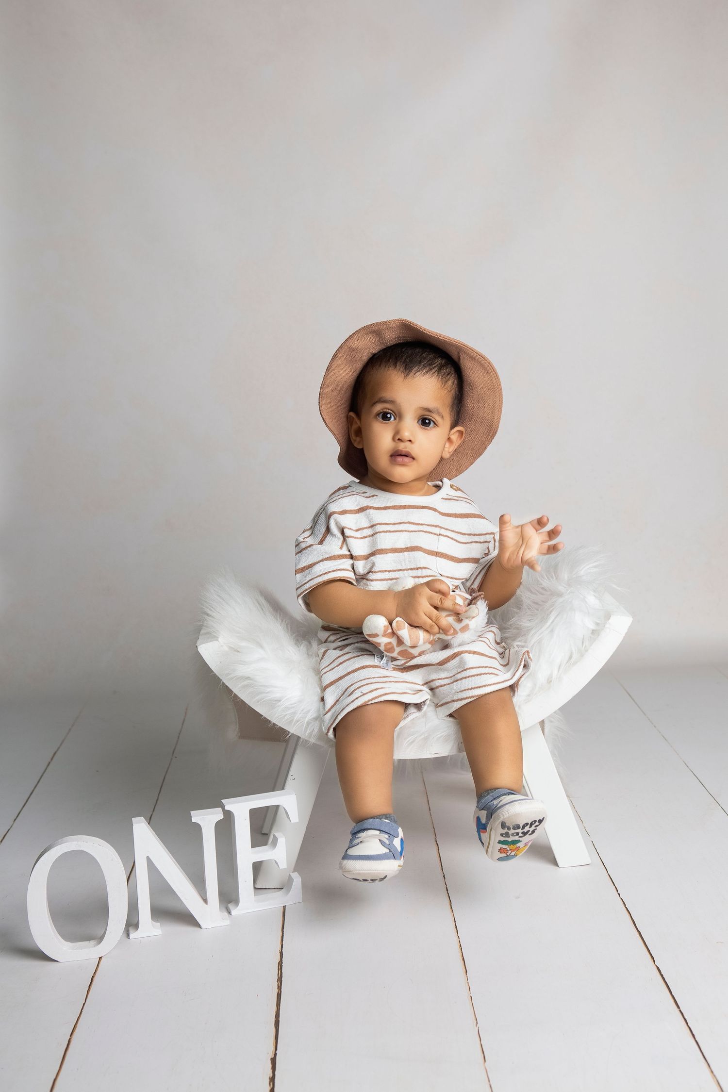 Adorable baby wearing a tan hat and striped outfit sits beside a white ONE sign celebrating their first birthday.
