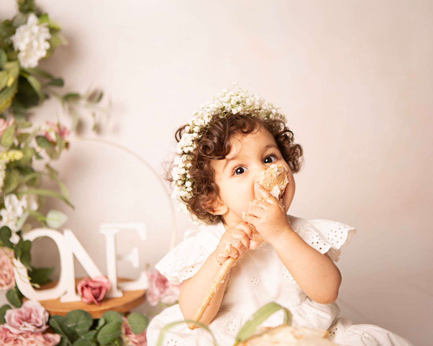 Toddler girl in white dress and floral crown eating cake at a first birthday smash cake photo session.