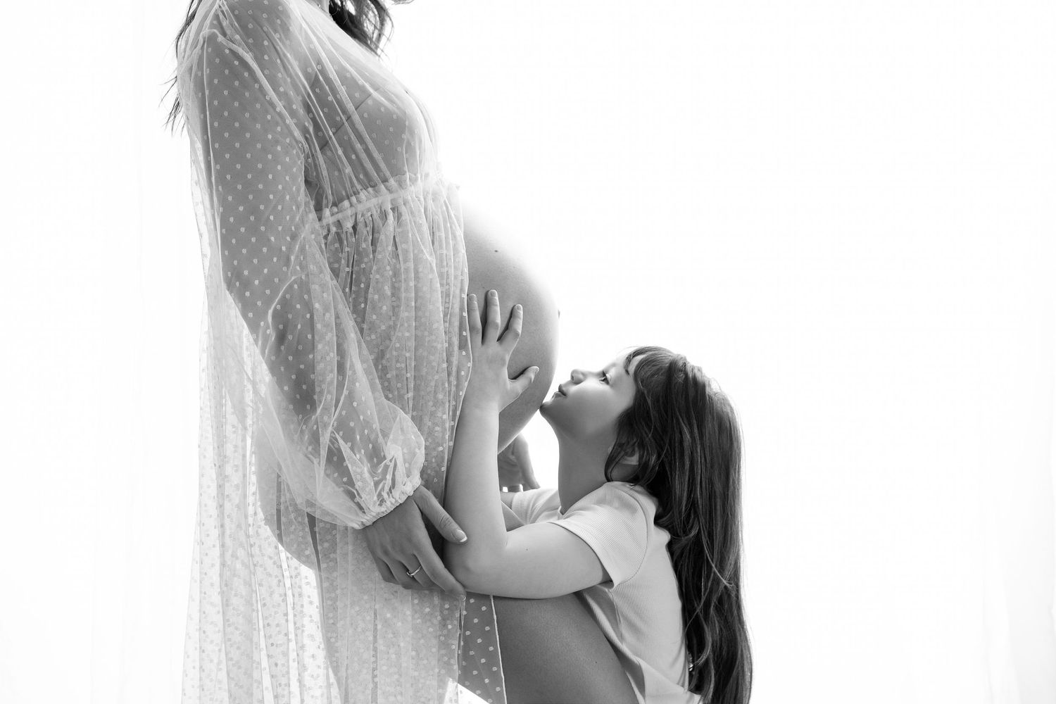 Black and white maternity photo of pregnant woman in lace gown with young girl kissing her baby bump.