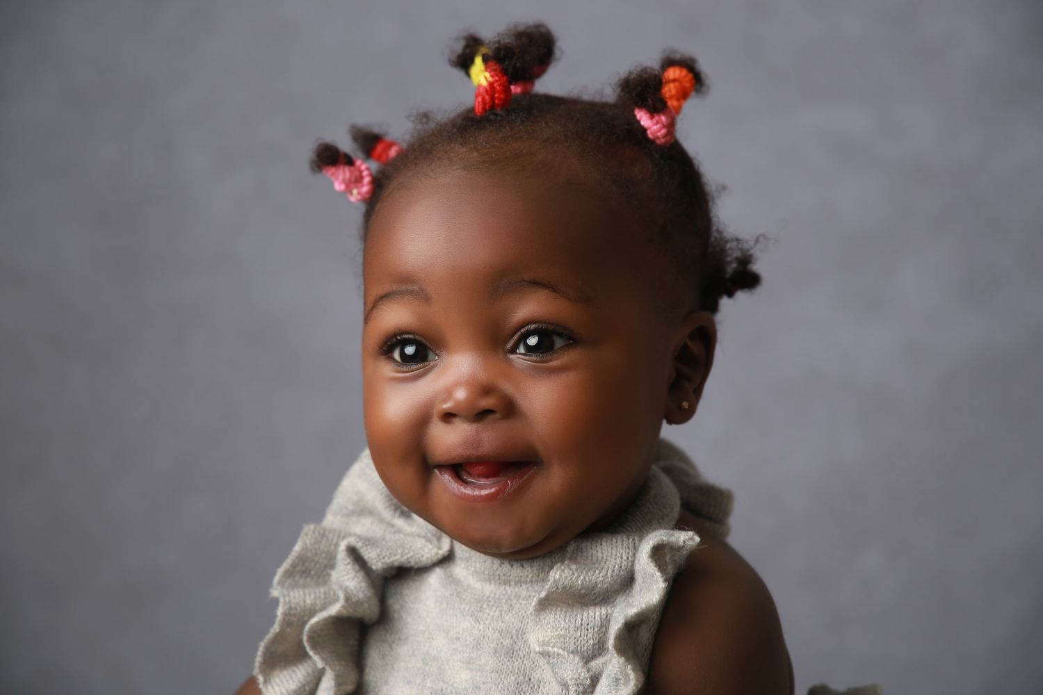 Smiling Black baby girl with colorful hair ties in tiny pigtails, wearing a gray ruffle outfit against a gray background.