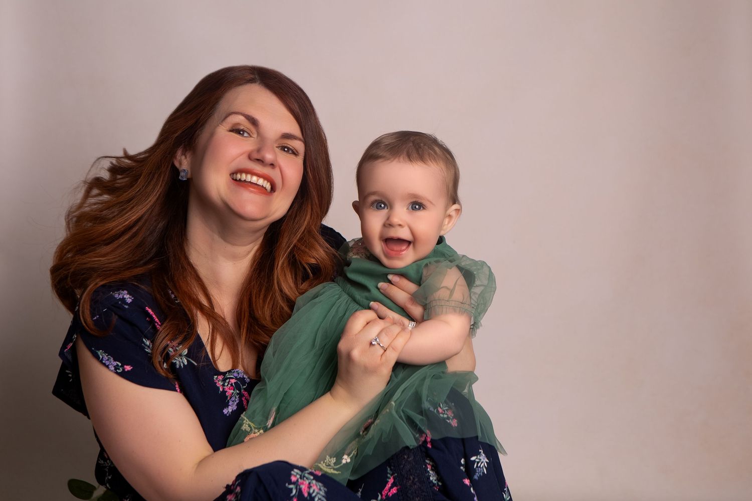 A joyful mother with red hair holds her laughing baby dressed in green, posing together for a studio portrait.