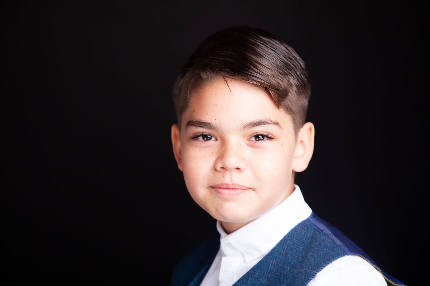 Young boy in a blue vest and white shirt smiling confidently against a dark background.