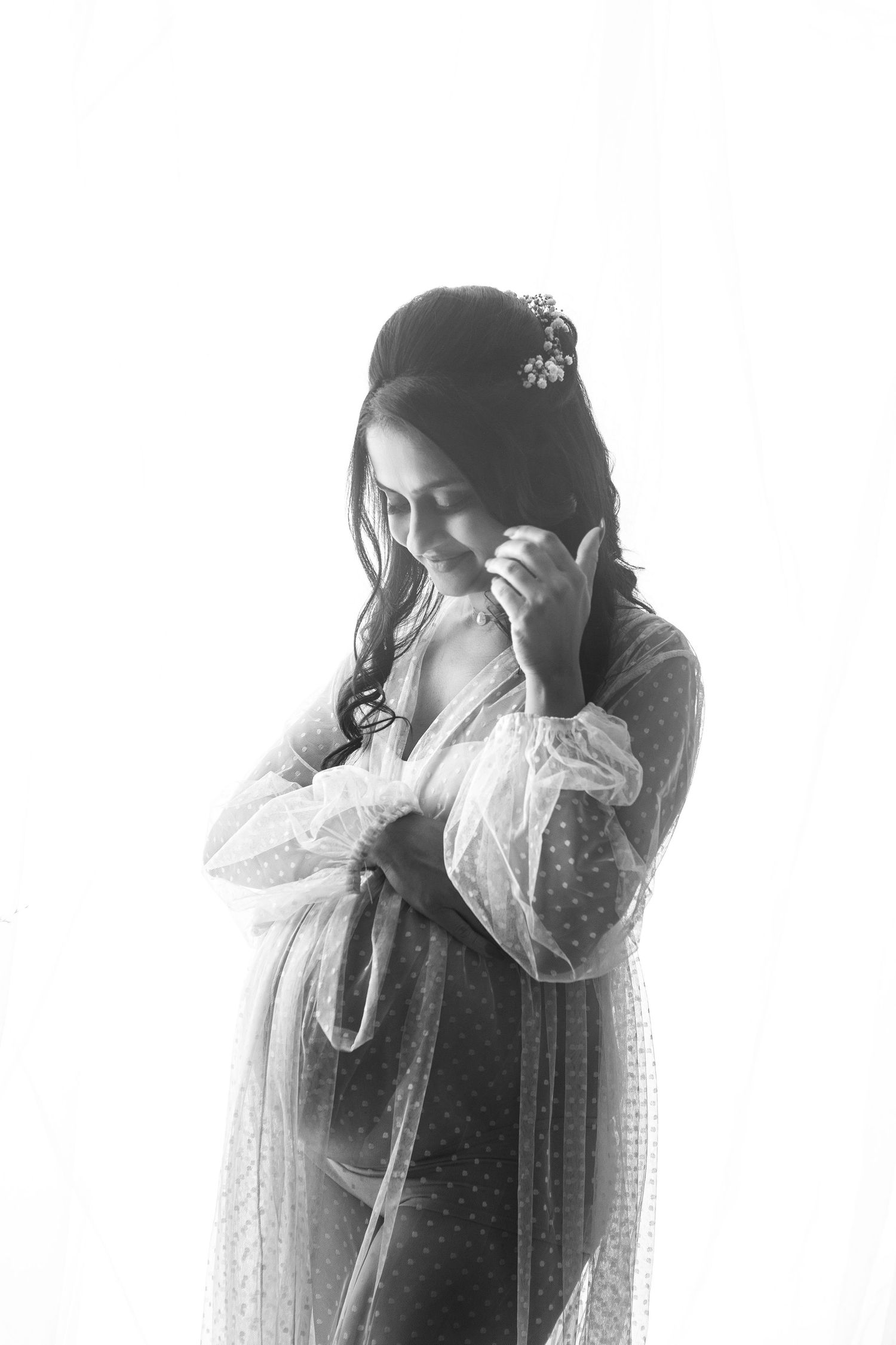 Black and white portrait of pregnant woman in sheer robe with floral hair accessory looking down.