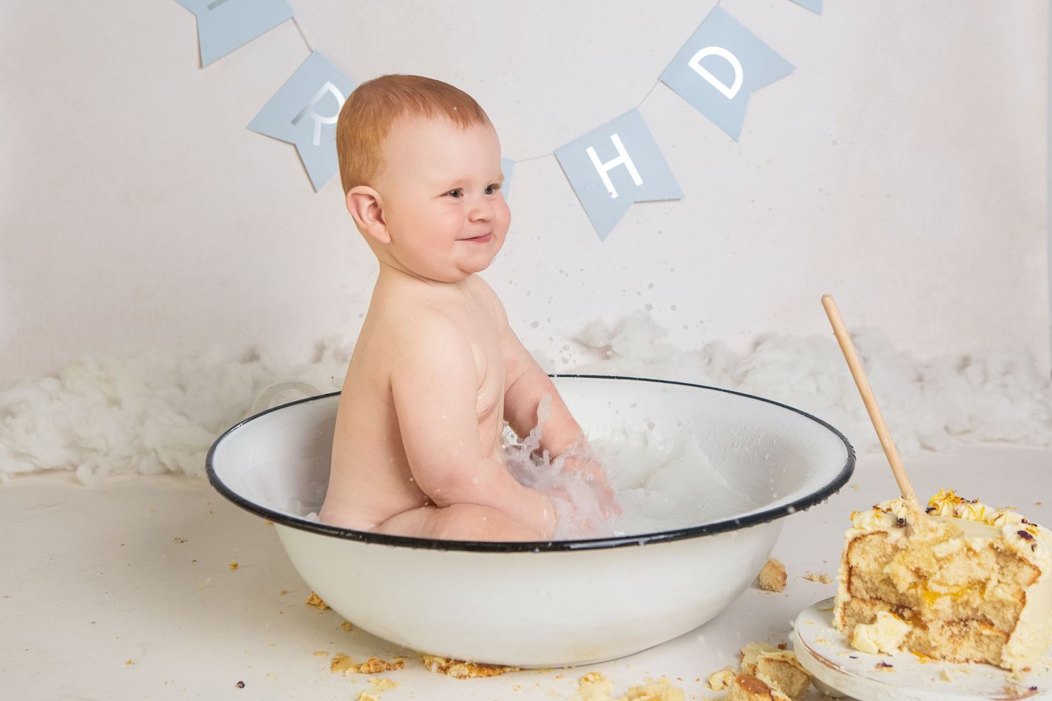 Baby boy sitting in vintage enamel tub during first birthday cake smash photo session with blue bunting.