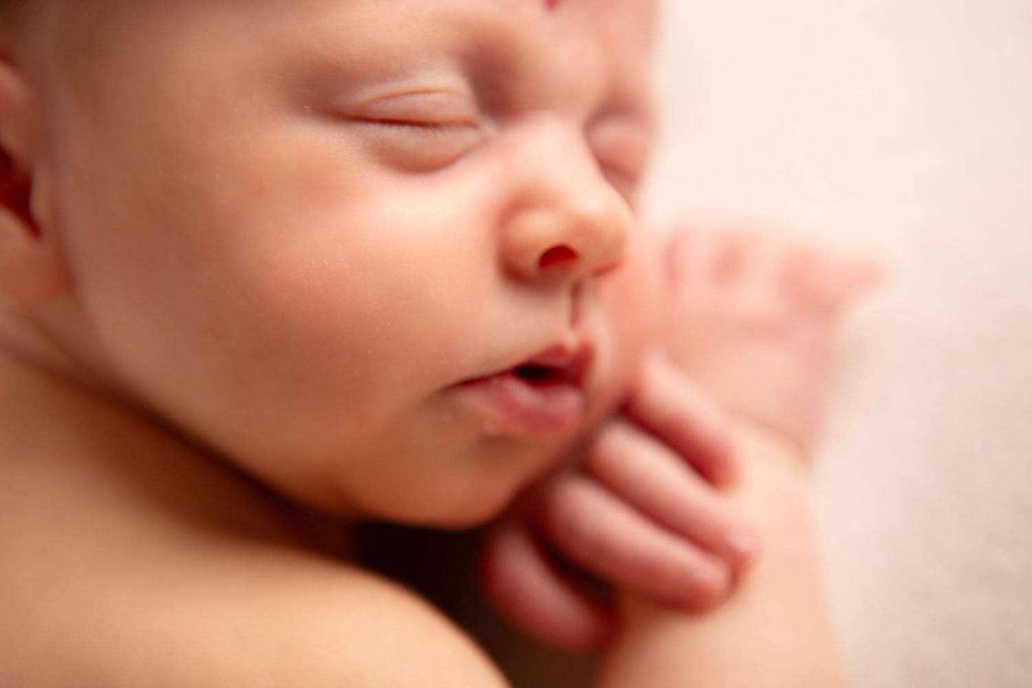 Close-up of a peaceful sleeping newborn baby with tiny hands tucked under chin on soft pink background.