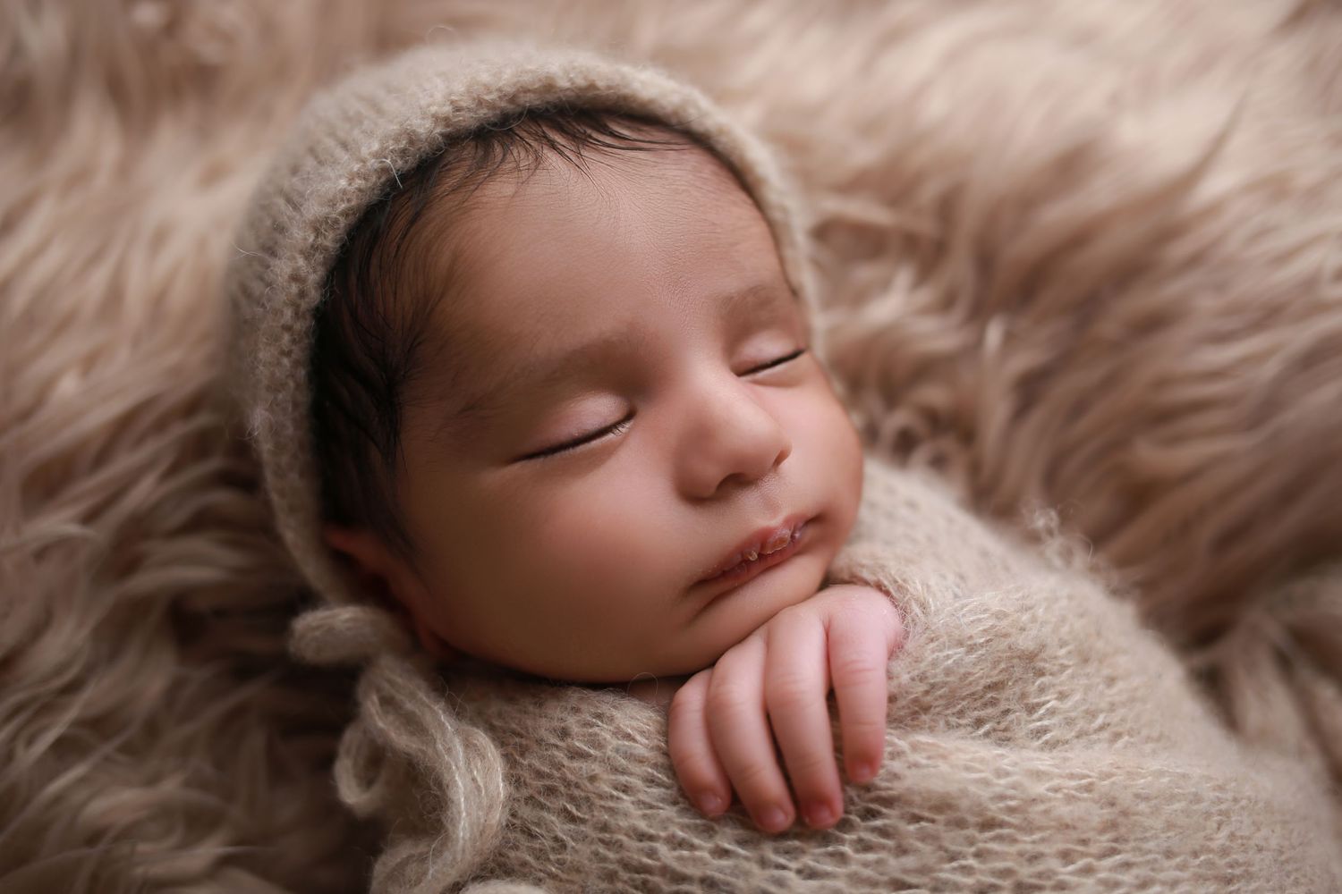 Peaceful newborn baby sleeping wrapped in a soft beige knit blanket and bonnet on a fluffy fur backdrop.