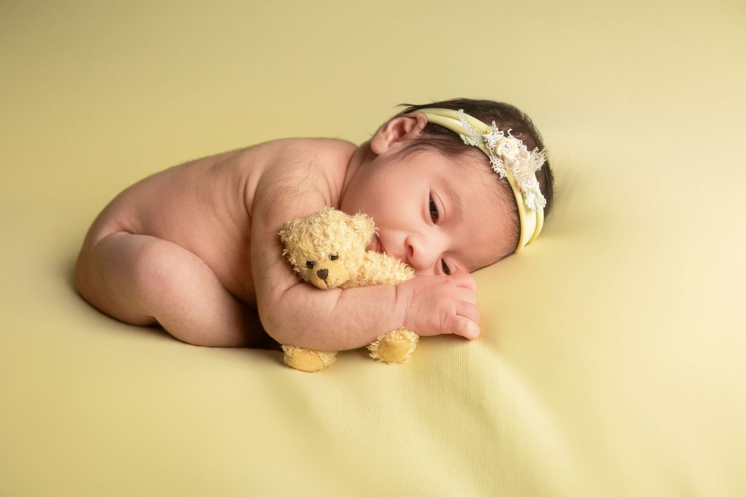 Sleeping newborn baby girl in yellow headband cuddling a small teddy bear on a soft yellow blanket.