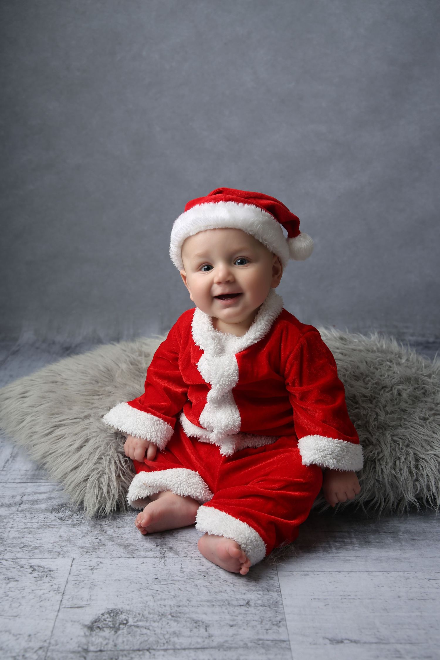 Adorable baby in red and white Santa Claus costume and hat sitting on fluffy rug for Christmas photo.