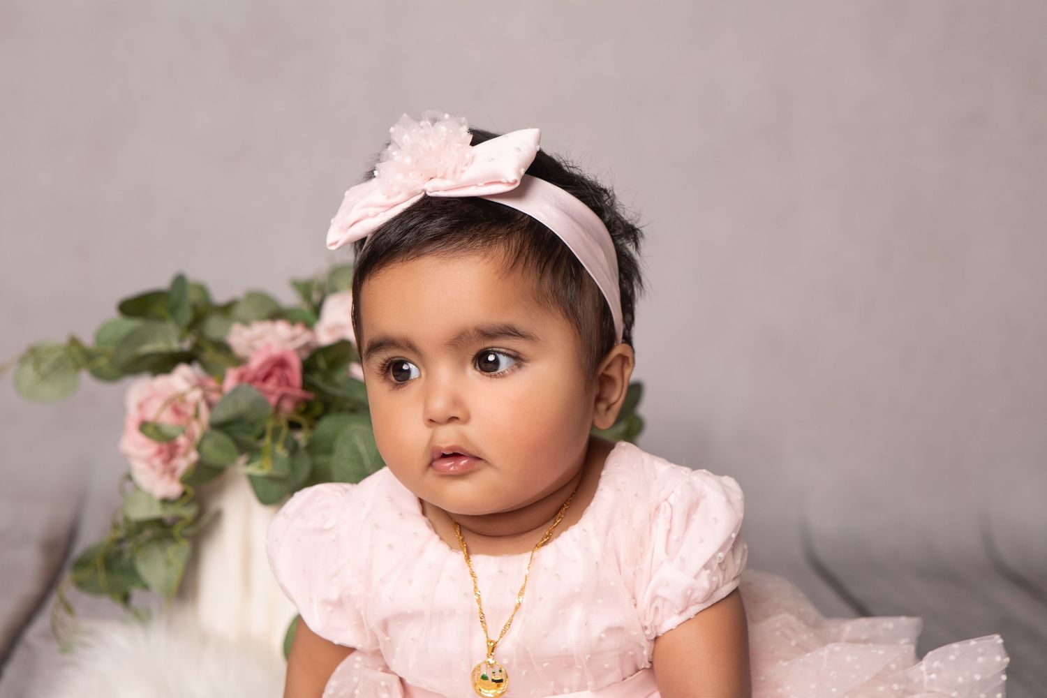 Adorable baby girl in pink dress and bow headband with gold necklace poses for portrait with floral backdrop.