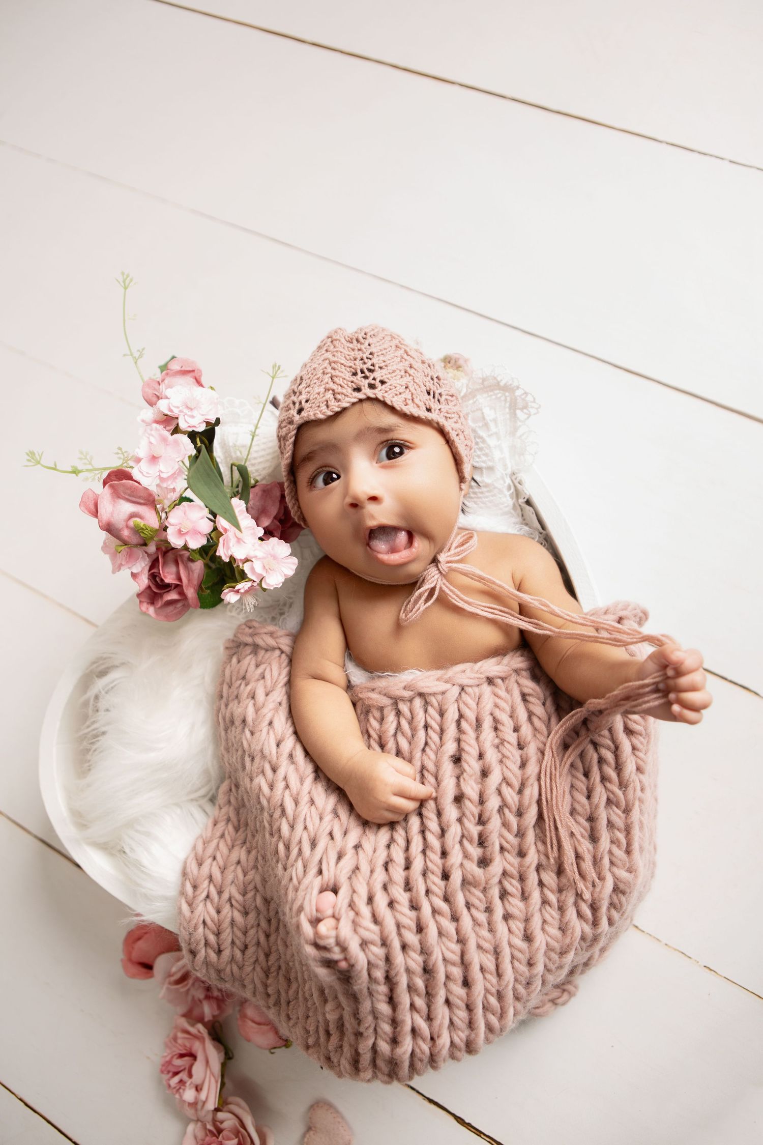 A funny, candid newborn moment of baby girl appearing to pull off her hat, surrounded by dusty pink tones.