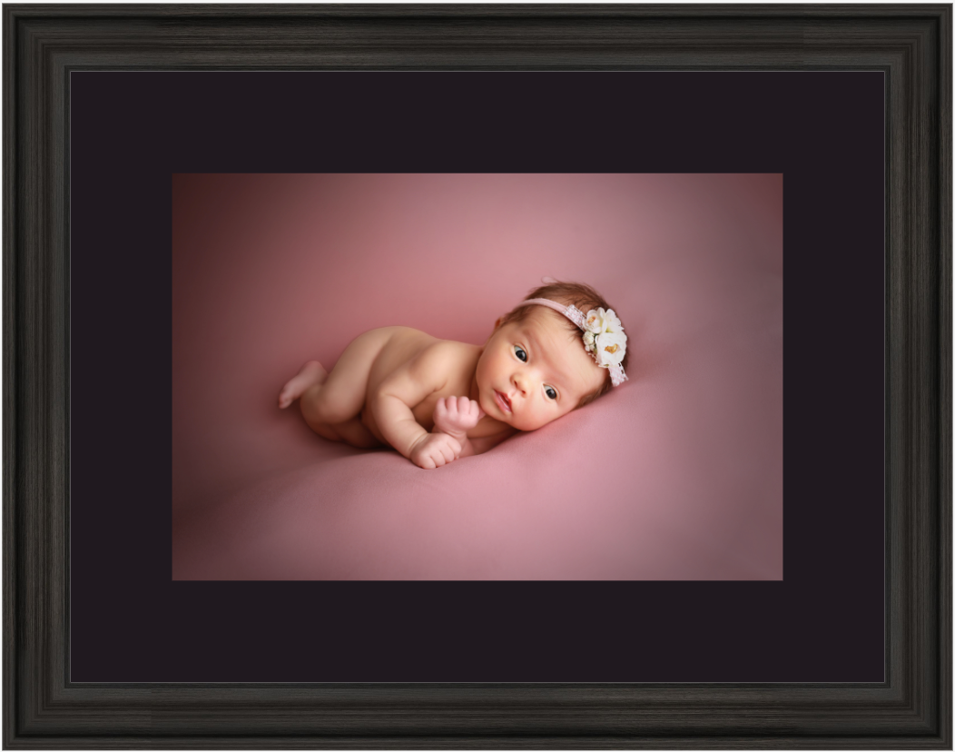 Newborn baby girl lying on pink backdrop wearing floral headband, displayed in dark wood frame with black matting.