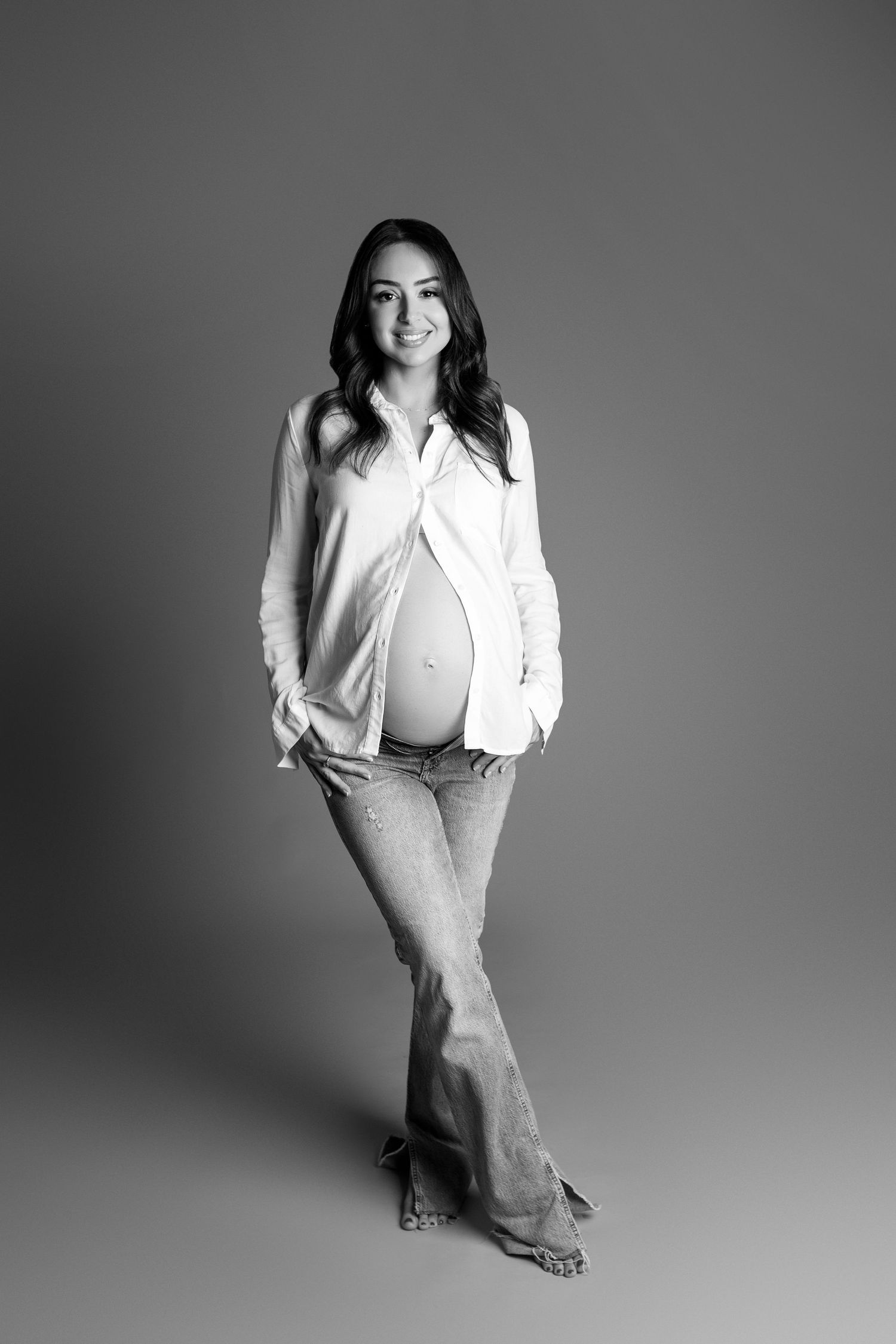 Black and white portrait of a smiling pregnant woman in open white shirt and flared jeans showing baby bump.