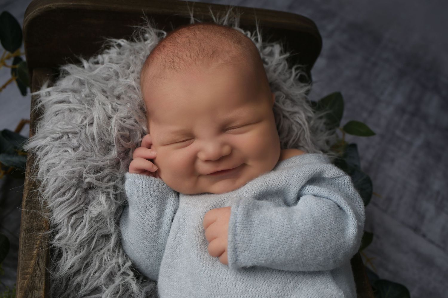 Smiling newborn baby in light blue onesie sleeping peacefully on soft gray fluffy blanket in wooden basket.