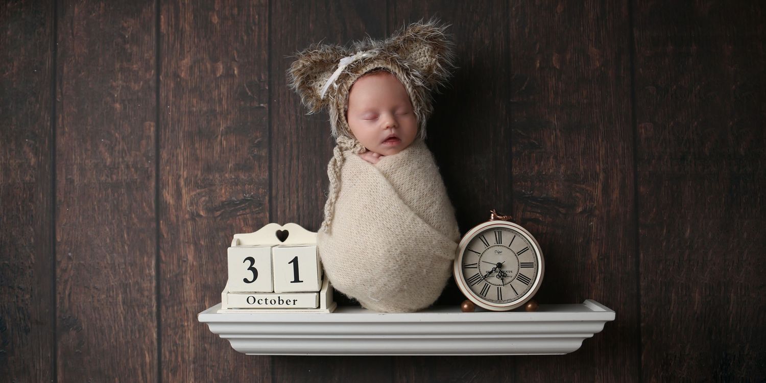 Newborn baby swaddled in cream wrap with bear bonnet, sleeping on shelf with October 31 calendar block and clock.