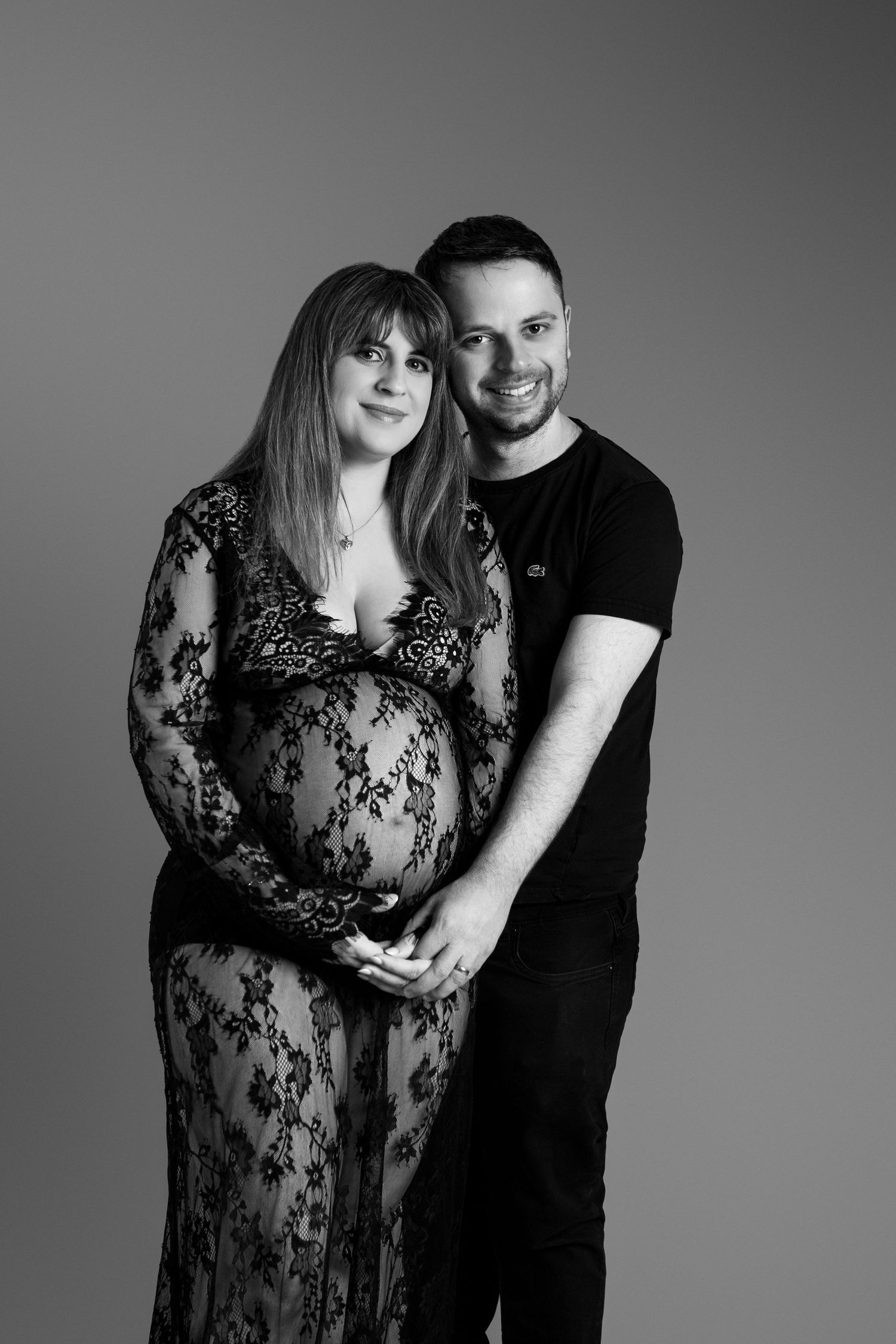 Black and white studio maternity portrait of couple, woman in floral lace gown, both smiling.