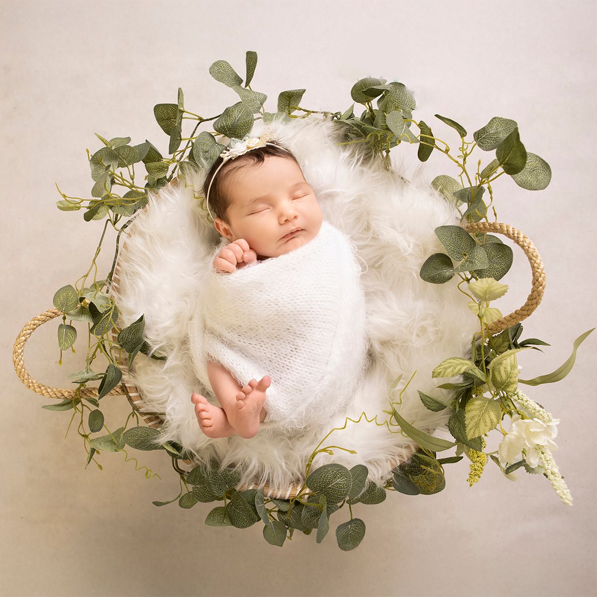 Sleeping newborn baby wrapped in white swaddled in a wicker basket with eucalyptus greenery and fluffy white fur.