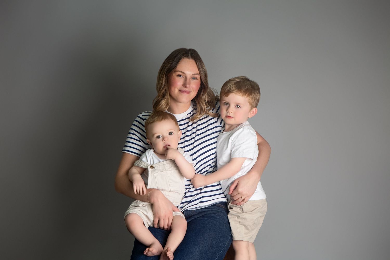 A woman in a striped shirt holds a baby and hugs a young boy during a professional family portrait session.