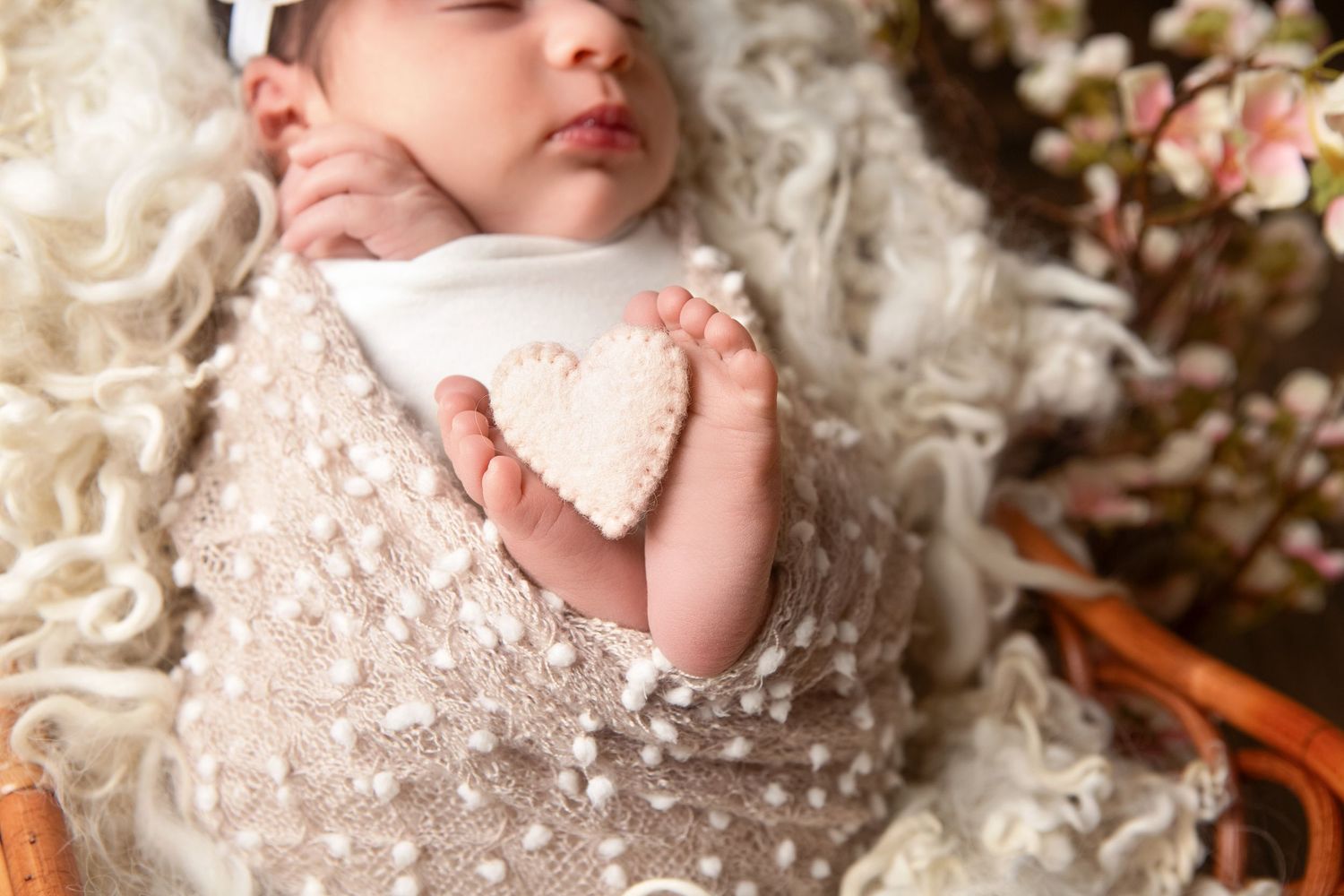 Newborn baby swaddled in a lacy beige wrap holding a small heart, nestled in a cozy basket with flowers.