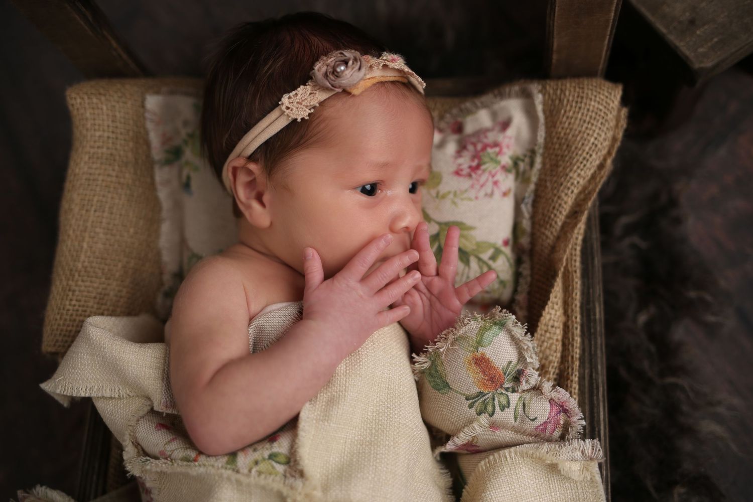 Newborn baby girl wearing a floral headband, resting in a vintage chair with hands near face.