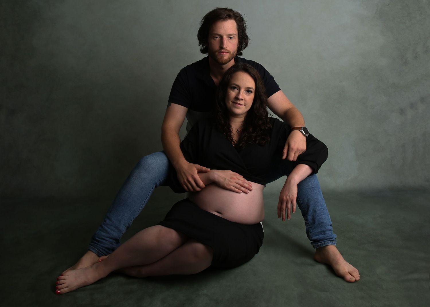 Couple poses for maternity photo shoot, woman in black dress showing baby bump, man in black shirt and jeans.