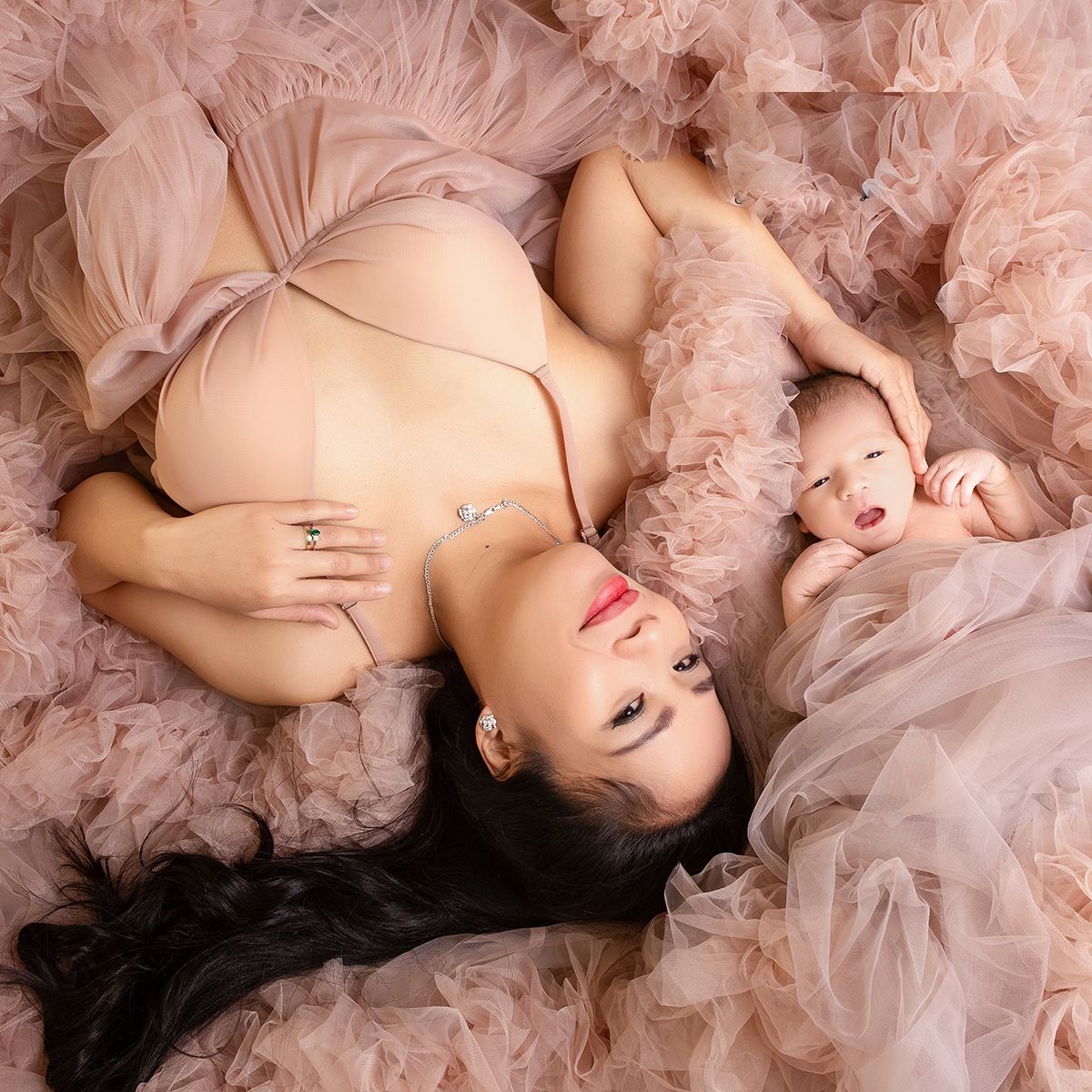 A pregnant woman in a blush pink gown lies beside her newborn baby on soft pink tulle fabric.