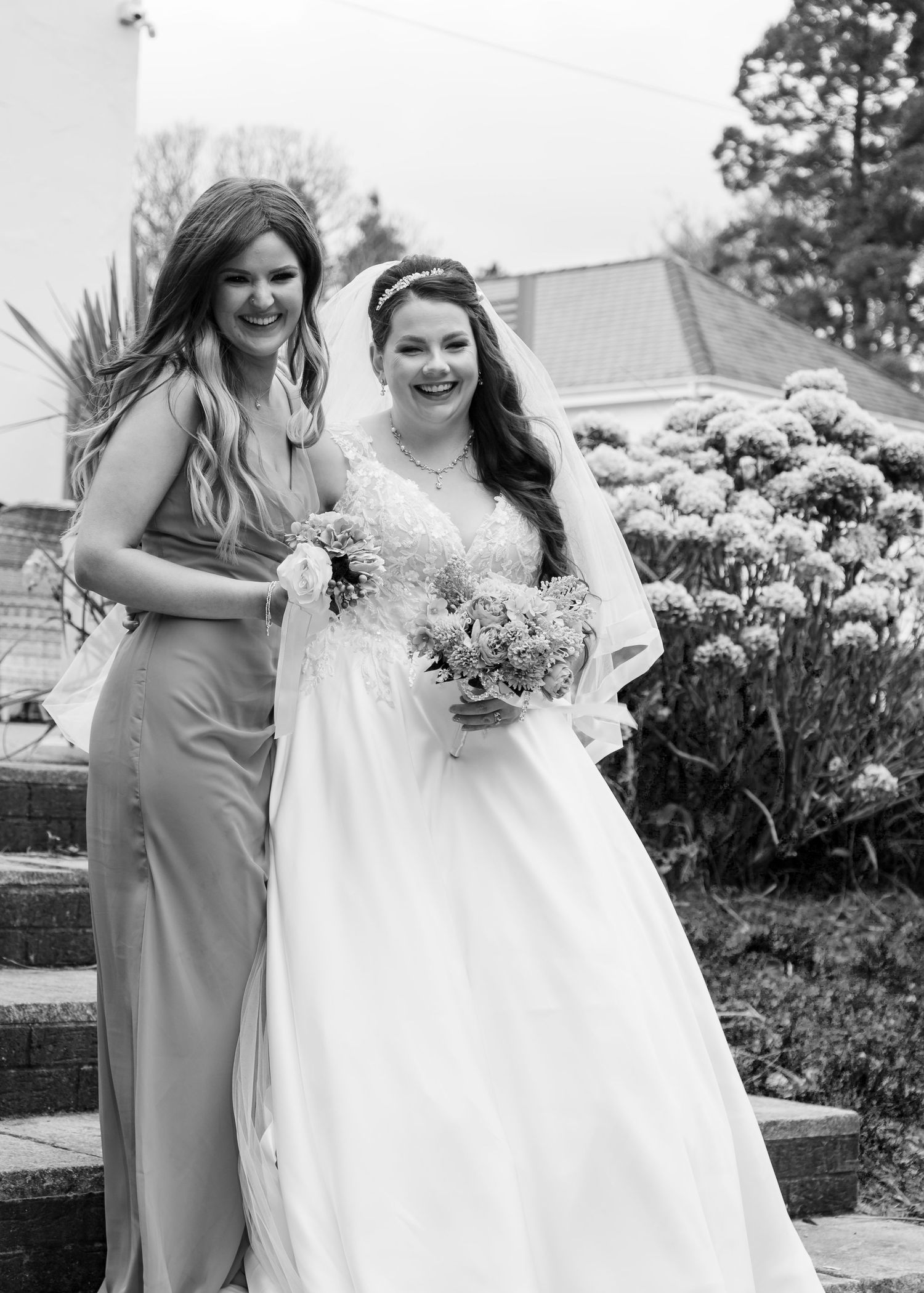 Black and white photo of a smiling bride in a white gown and veil holding a bouquet, standing with her bridesmaid outdoors.