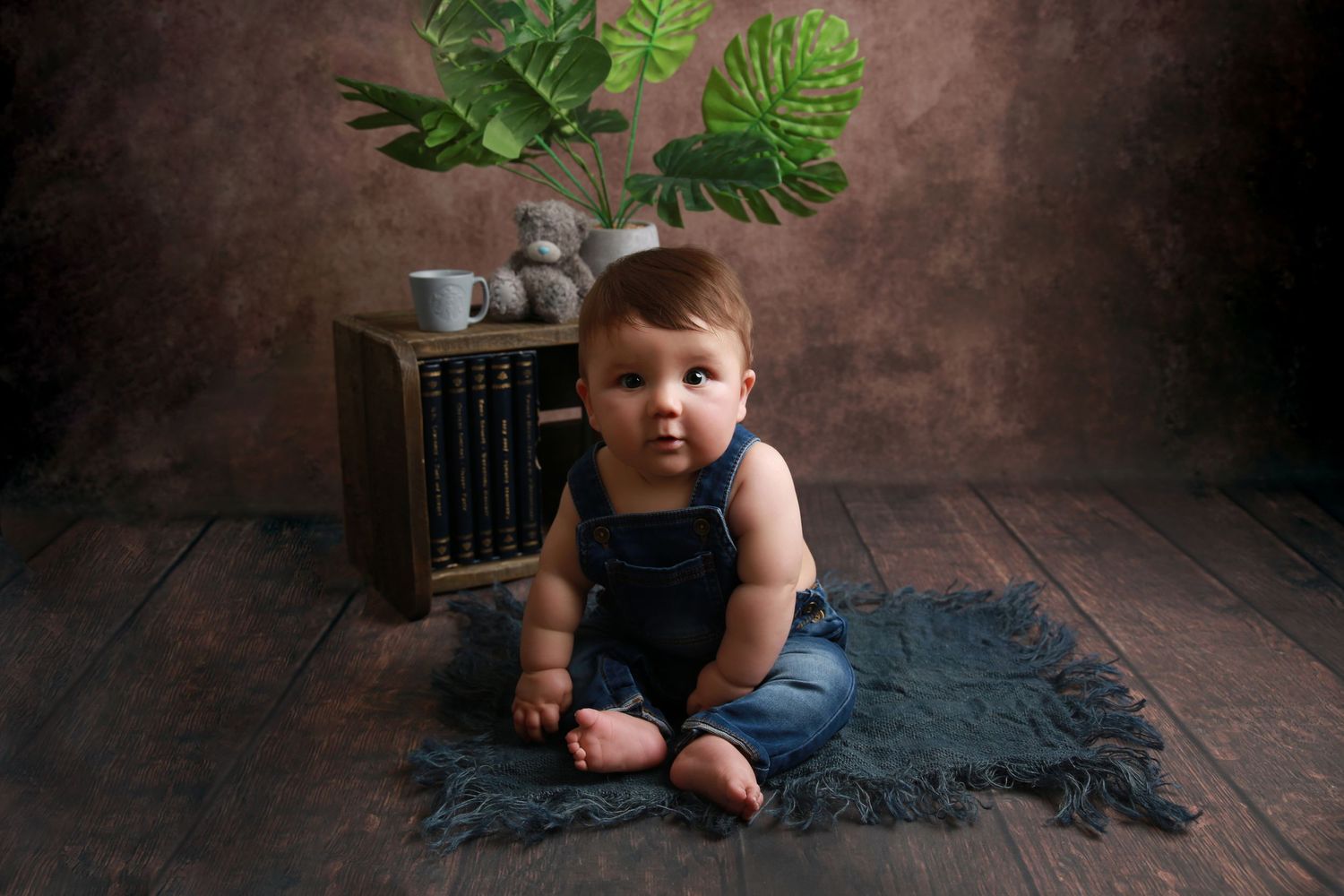 A baby in denim overalls sits on a blue rug with a wooden crate, teddy bear, and tropical leaf backdrop.