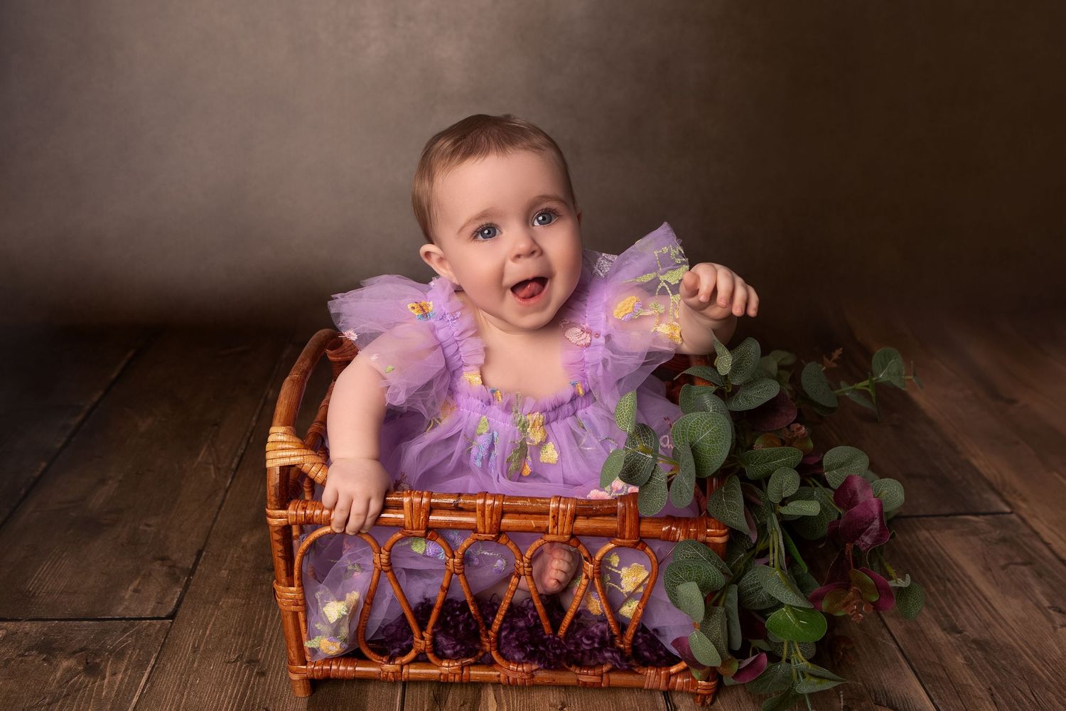 Baby girl in purple tutu dress sits smiling in wicker basket with greenery on wood floor.