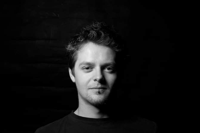 Black and white portrait of a young man with curly hair wearing a dark shirt against a dark background.