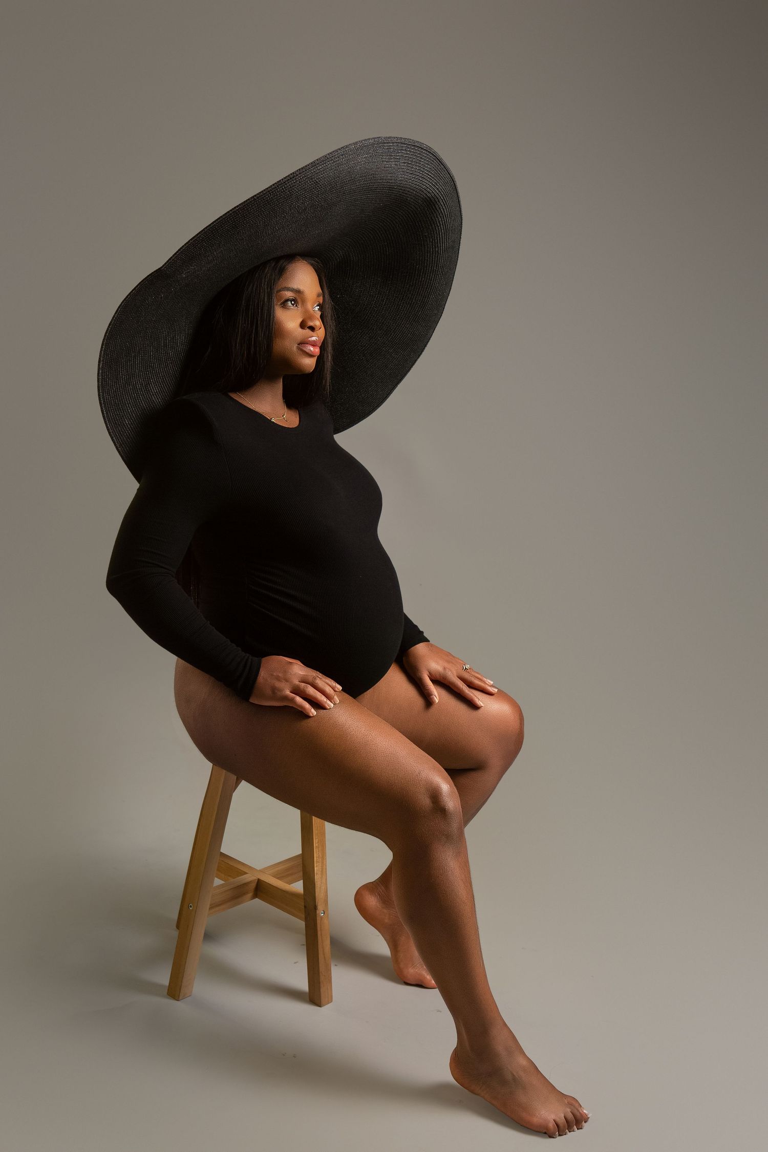 Pregnant Black woman in black bodysuit and wide-brim hat seated on wooden stool.