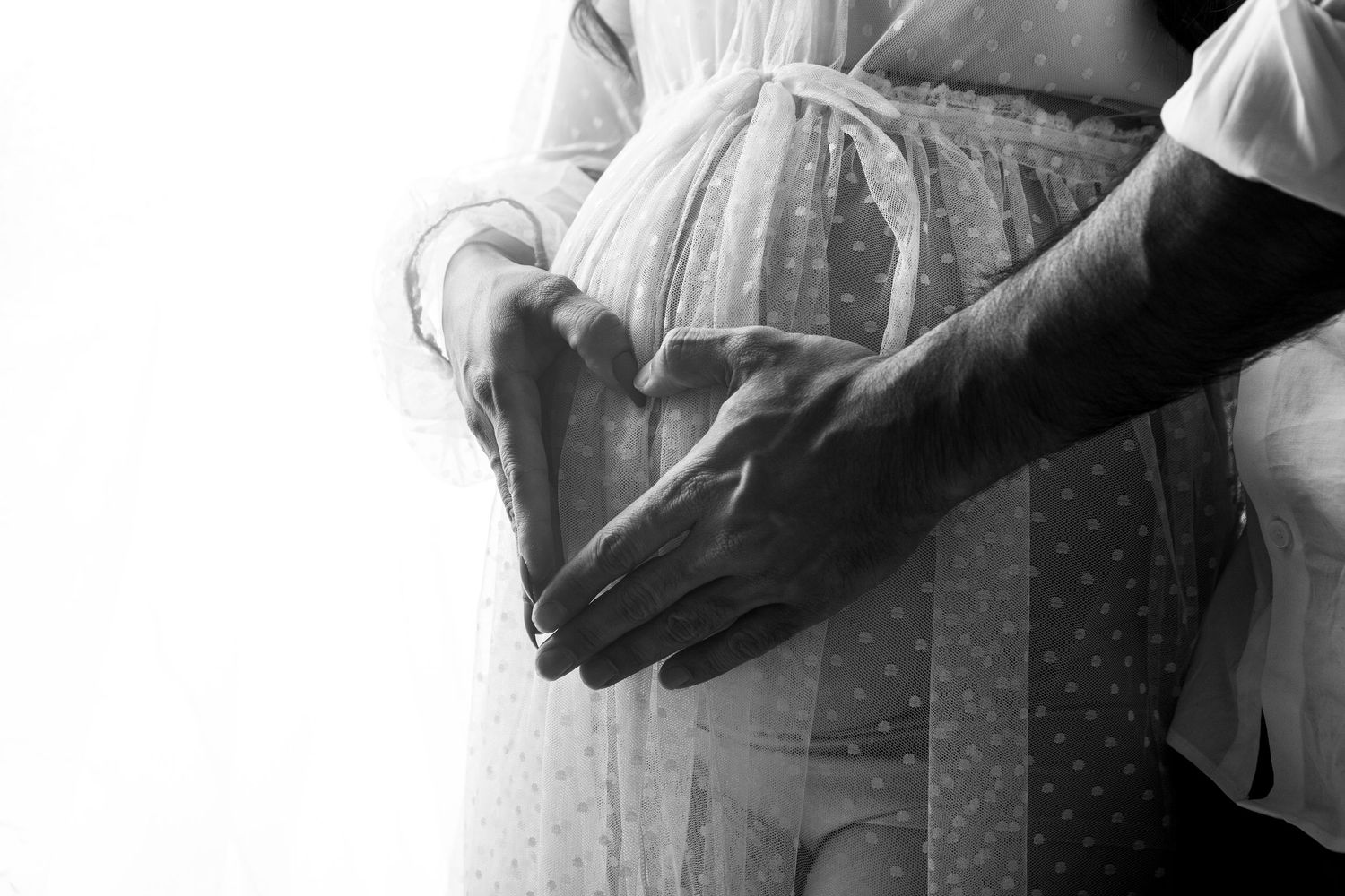 Black and white close-up of couple forming heart shape with hands over pregnant belly.