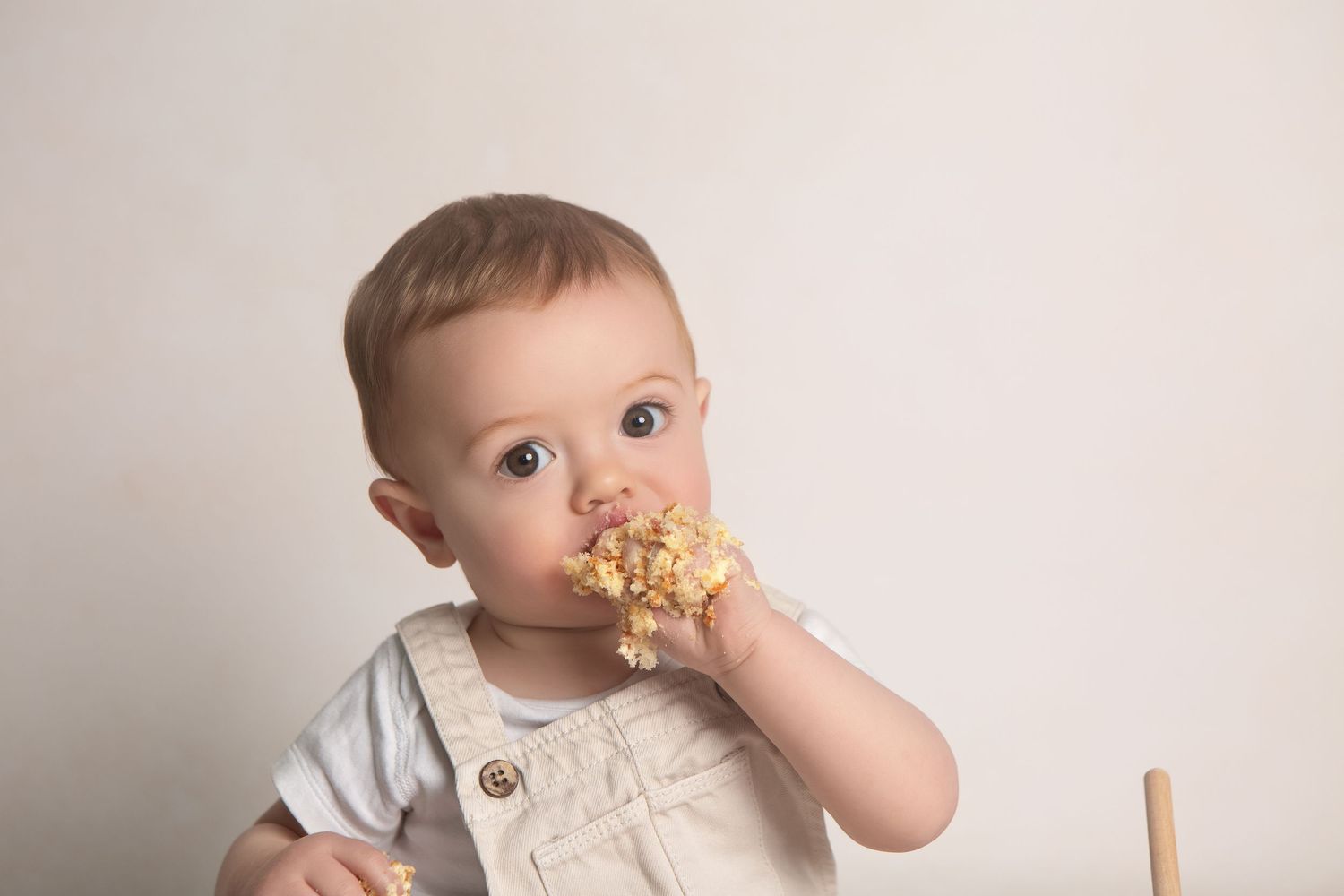 Cute baby in overalls eating a smash cake with both hands, celebrating their first birthday.