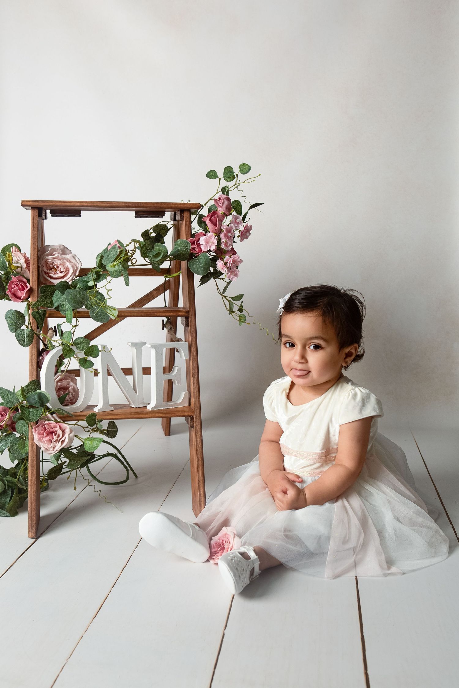 A baby girl in a white tutu dress sits on a white floor beside a wooden ladder decorated with pink roses and ONE sign.