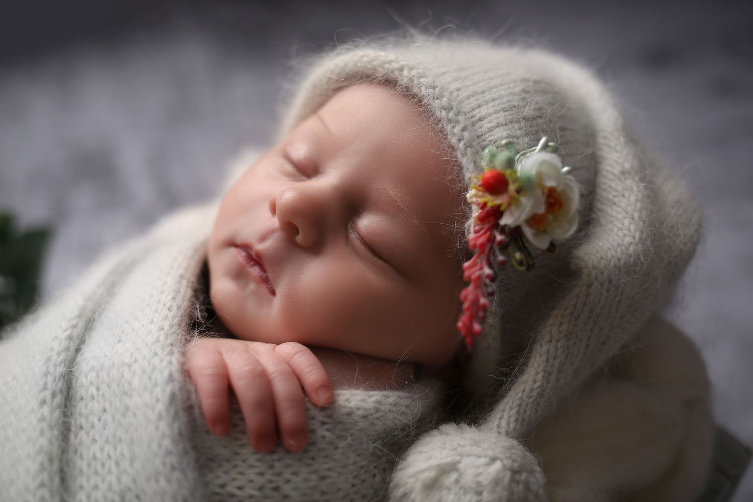 Sleeping newborn baby wrapped in soft gray knit blanket wearing a cozy bonnet adorned with delicate flowers.