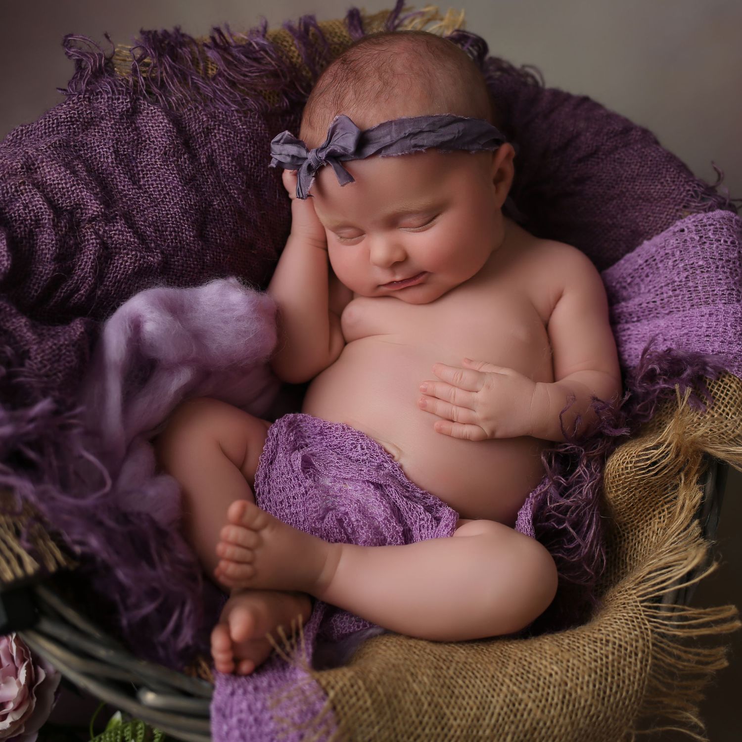 Newborn baby girl sleeping peacefully in a purple knit basket with soft lavender wraps and a bow headband.