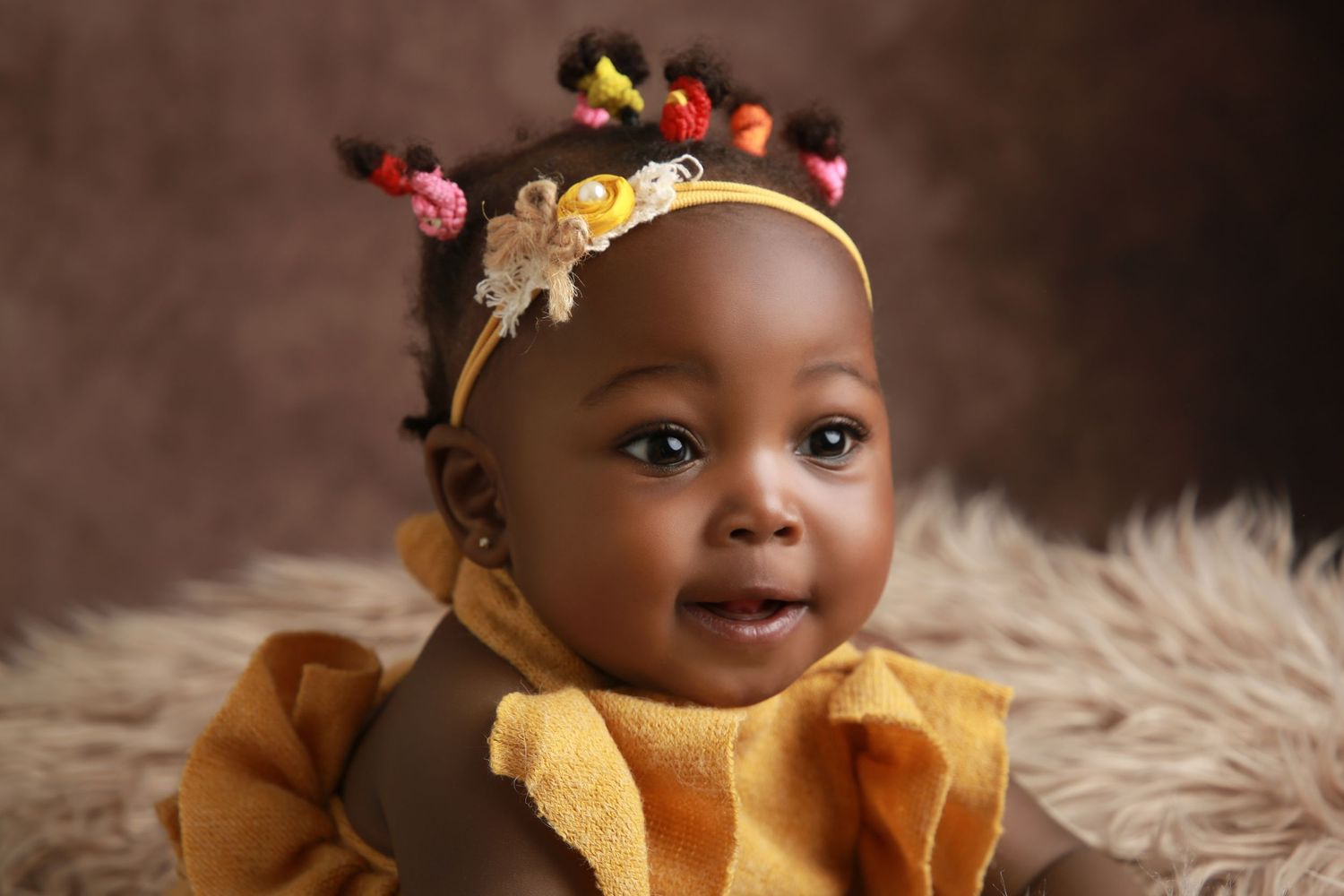 Adorable baby girl in yellow ruffled outfit with colorful hair accessories and headband, smiling at camera.