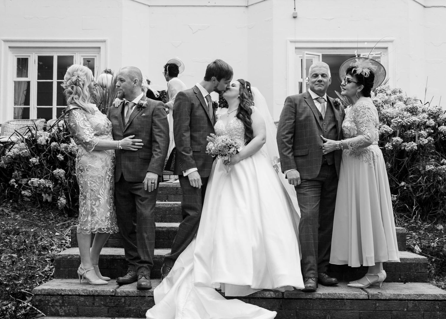 Black and white wedding photo of bride and groom kissing surrounded by family on outdoor steps.