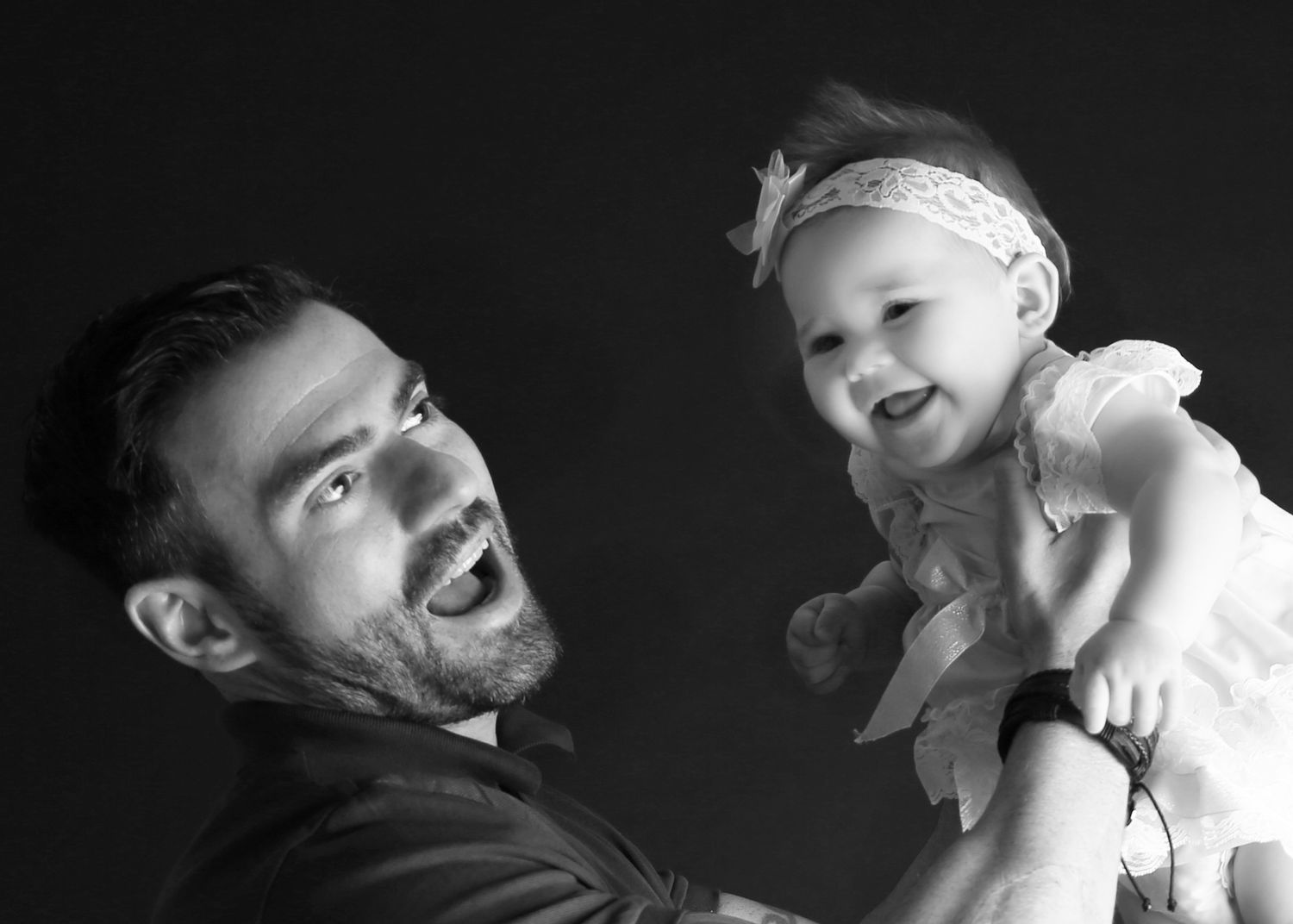Black and white photo of a smiling man holding a laughing baby girl wearing a white dress and headband.