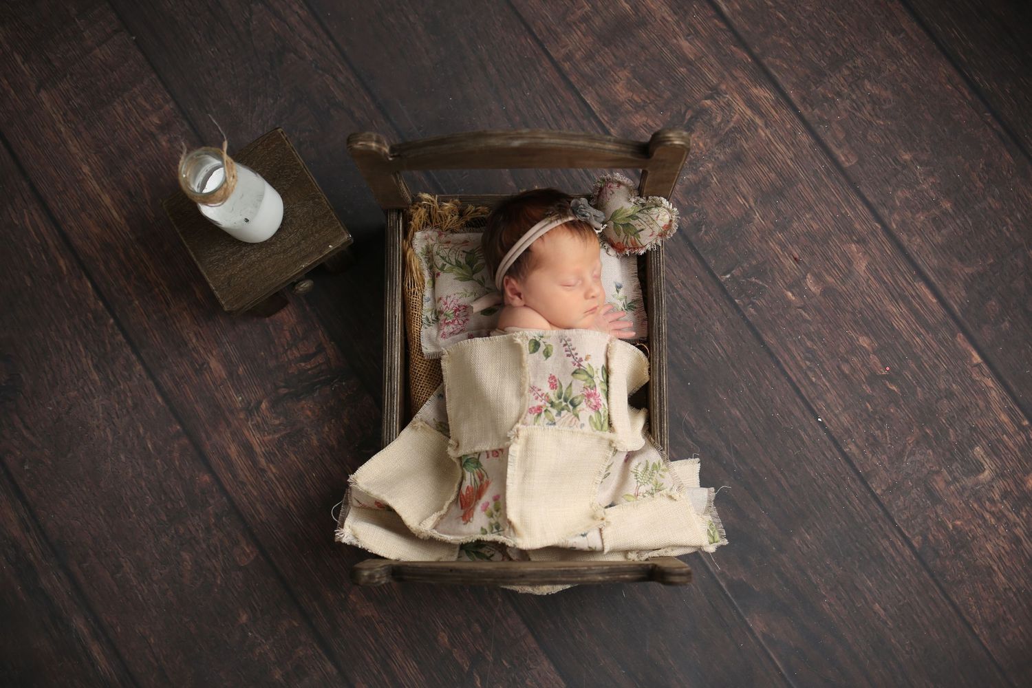 Newborn baby girl sleeping in a tiny vintage wooden bed wearing a floral gown and headband.