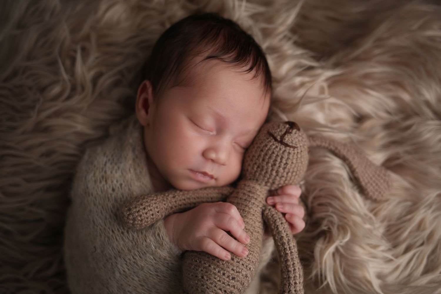 Sleeping newborn baby wrapped in a cozy knit blanket, cuddling a small teddy bear on a fluffy beige rug.