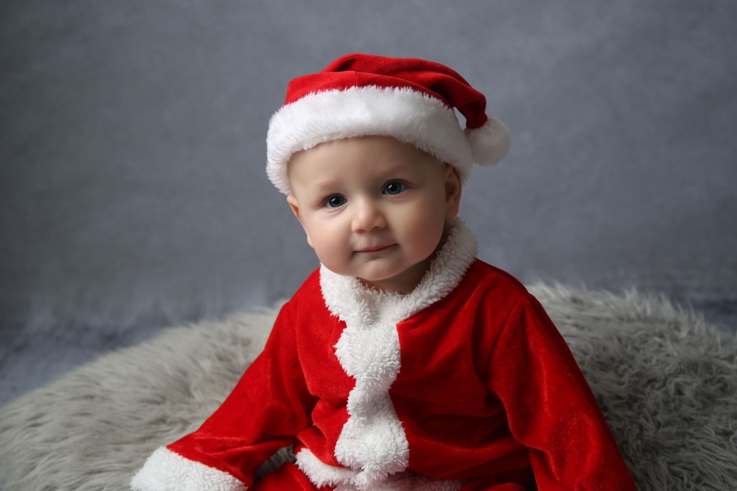 Adorable baby dressed in a red Santa Claus outfit and hat, smiling against a gray background.