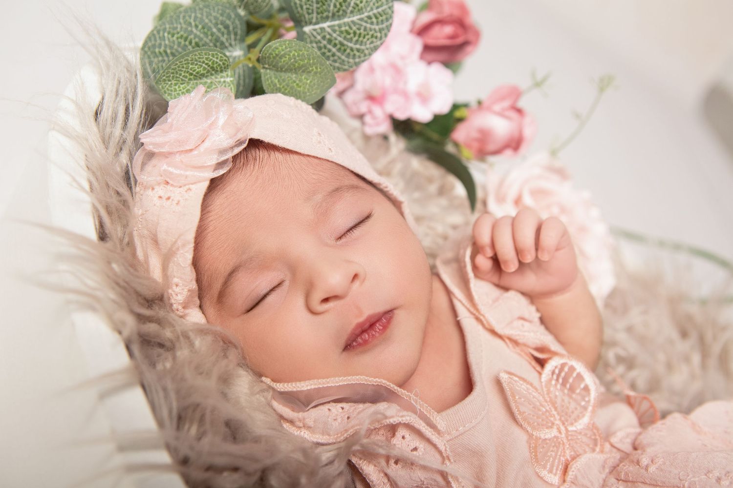 Sleeping newborn baby in pink outfit and floral headband nestled in a fluffy basket with roses.