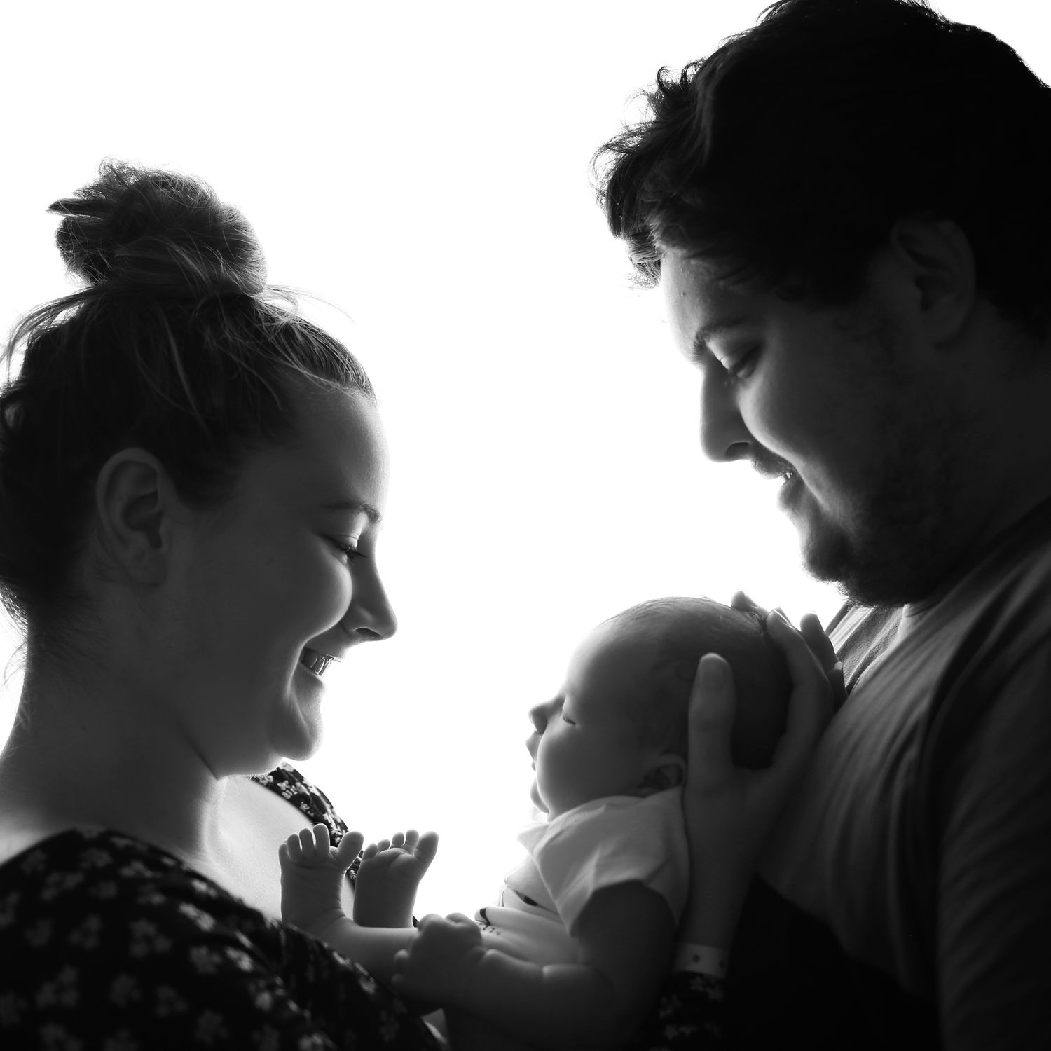 Black and white photo of a couple smiling at their newborn baby, captured in a tender family portrait.